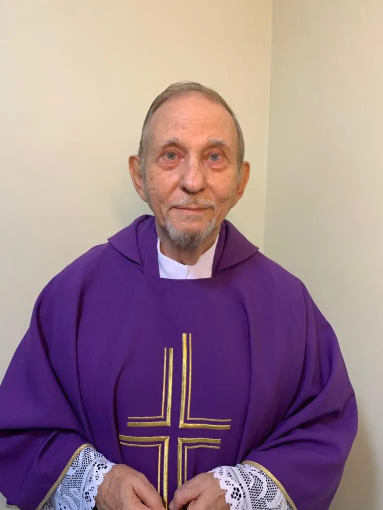 Pessoa idosa vestindo paramento religioso roxo com detalhe de cruz dourada no centro, vista de frente, com as mãos unidas à frente do corpo, diante de fundo neutro claro.