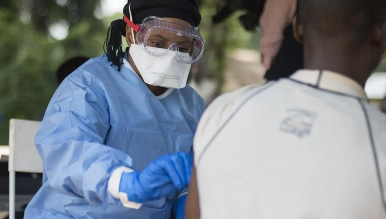 Enfermeira negra com roupa azul e viseira transparente aplica medicamento em outra pessoa.