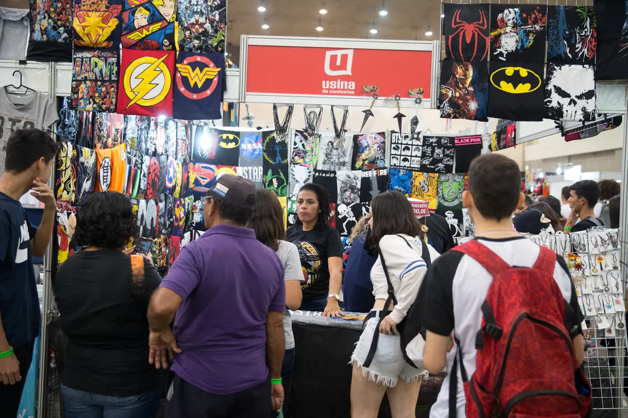 Frequentadores de um evento geek interagem em frente a um estande repleto de camisetas e acessórios de super-heróis, bandas e cultura pop, sob a placa vermelha da marca 'Usina de Camisetas'. A imagem captura o movimento do público de costas e de perfil, concentrado em olhar os produtos expostos em uma atmosfera movimentada e colorida.