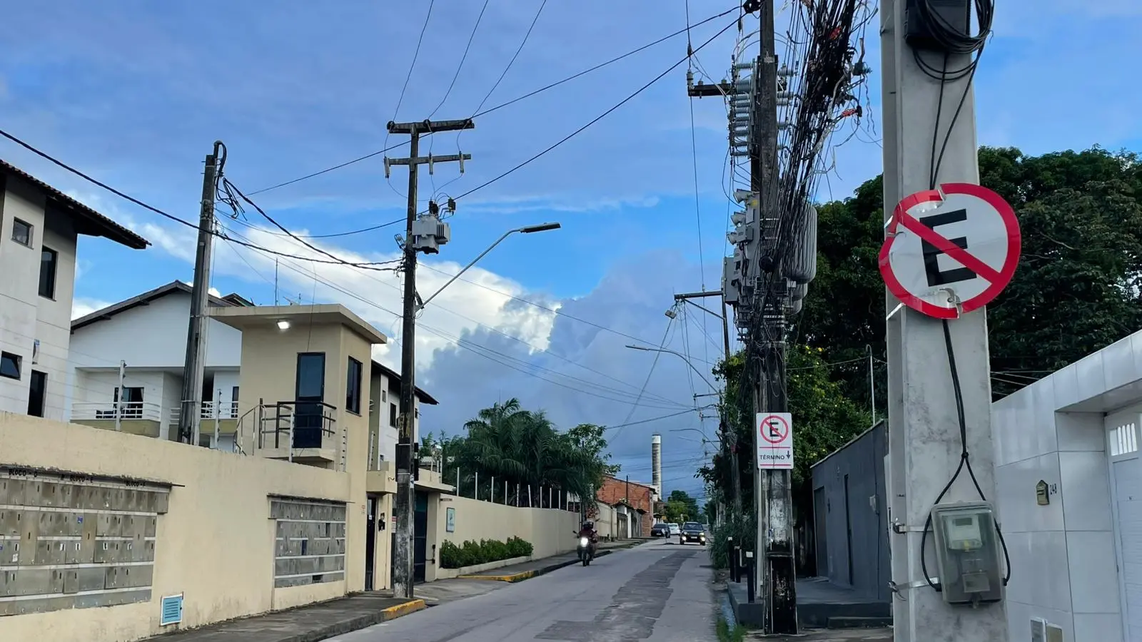 Rua ladeada por apartamentos e postos com fios de energia em Caucaia. Ao fundo, céu nublado com nuvens escuras.