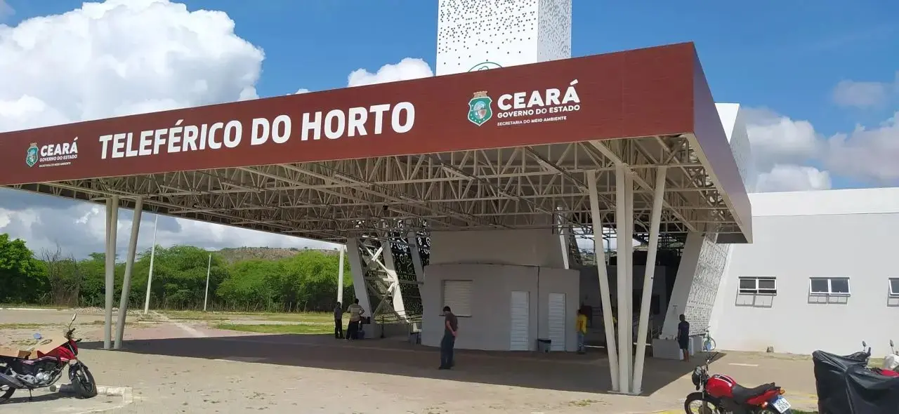 Plataforma do Teleférico do Horto, em Fortaleza, Ceará, com placa “Teleférico do Horto” e “Ceará Governo do Estado”, estrutura metálica e moto estacionadas ao lado, sob céu azul e nuvens.