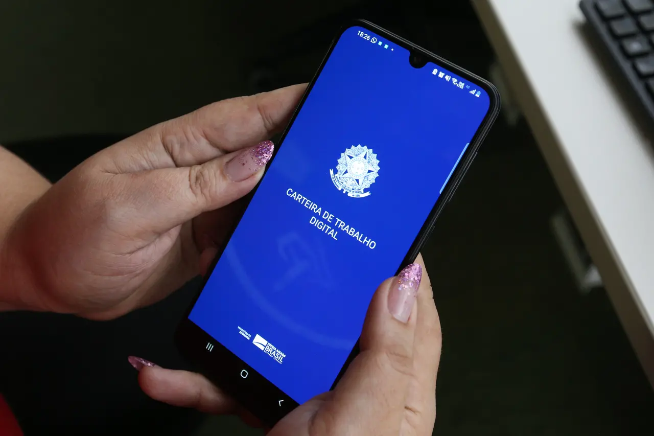 Mãos com unhas decoradas em glitter rosa seguram um smartphone que exibe a tela inicial azul do aplicativo Carteira de Trabalho Digital, com o brasão do Brasil em destaque. A imagem, capturada em ângulo diagonal, mostra o aparelho apontado para cima em um ambiente de escritório com parte de um teclado visível ao fundo.