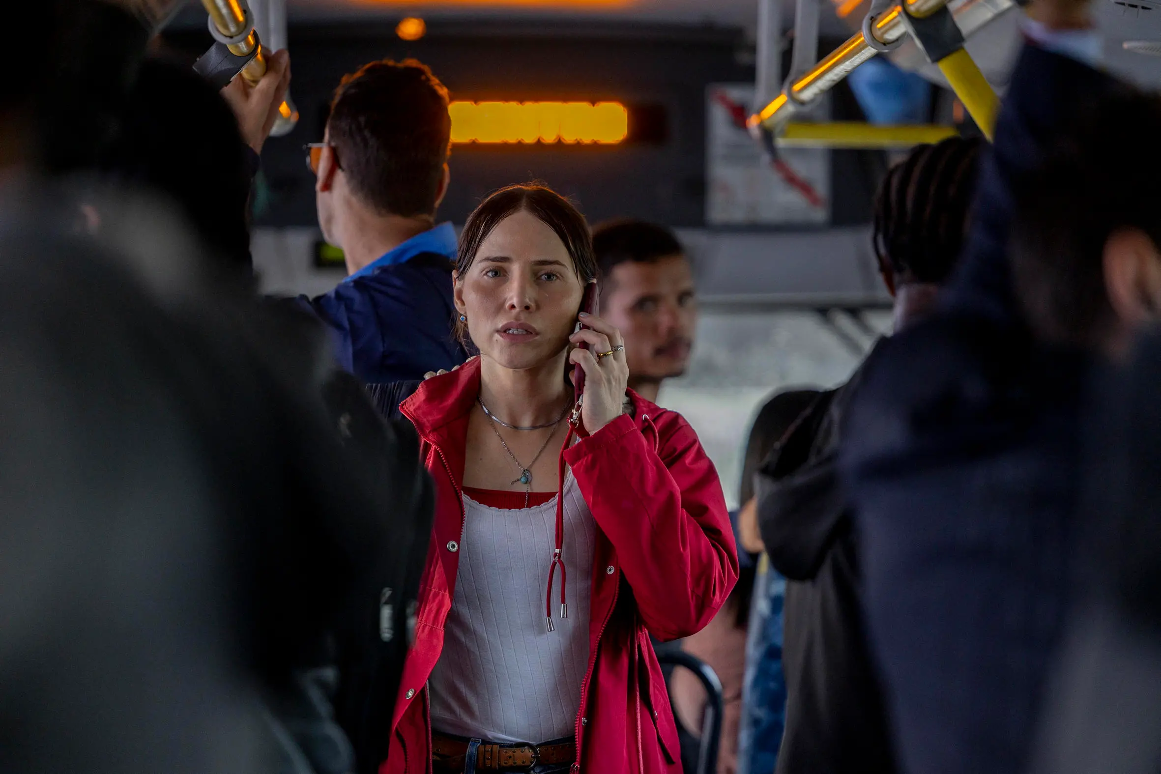 Pessoa em primeiro plano está dentro de um transporte público, segurando um celular junto ao ouvido, com braços erguidos para se equilibrar entre as barras superiores. Veste casaco vermelho sobre blusa clara. Ao redor, outras pessoas aparecem desfocadas, algumas também segurando os suportes do veículo. O ambiente interno tem iluminação mista, com luz natural entrando e pontos de luz artificiais ao fundo, criando contraste com o movimento e a proximidade entre os passageiros.
