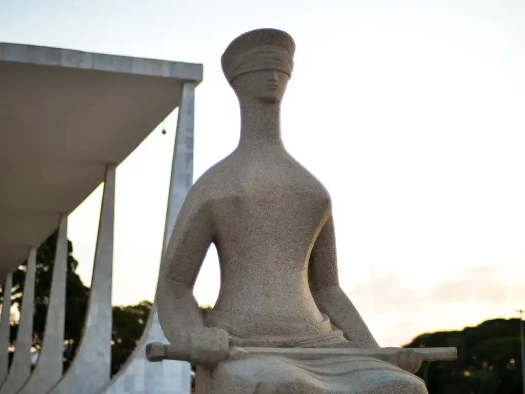 Estátua “A Justiça”, em frente ao Supremo Tribunal Federal, em Brasília. Escultura de pedra aparece sentada diante da fachada do STF ao entardecer.