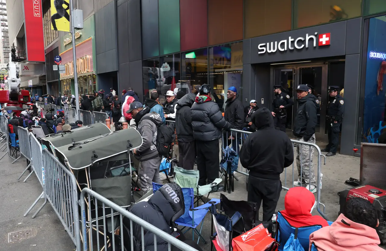 Uma multidão de pessoas faz fila e aguarda sentada em cadeiras de praia atrás de grades de metal na calçada em frente a uma loja da Swatch na Times Square, em Nova York. Policiais monitoram a entrada do estabelecimento enquanto os clientes, agasalhados com casacos de inverno e capuzes, se organizam de forma linear ao longo da rua movimentada.