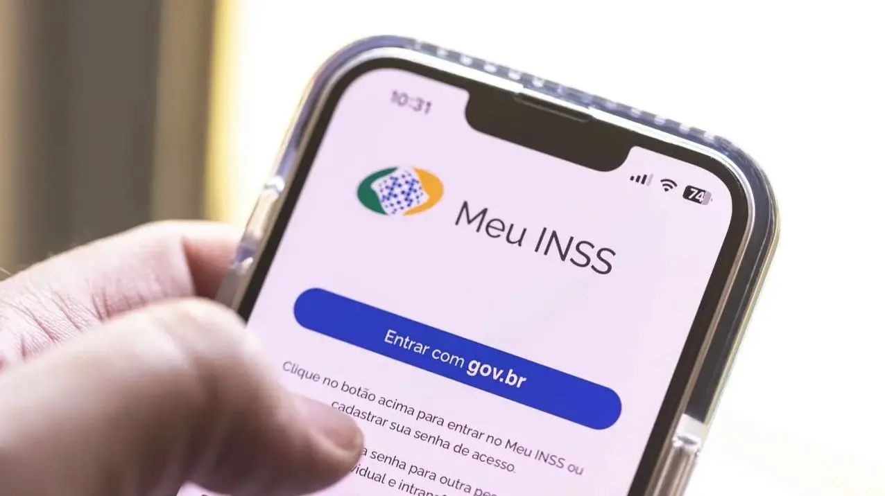 Celular exibindo a tela do aplicativo Meu INSS, com botão “Entrar com gov.br”, sendo segurado por uma mão em primeiro plano.