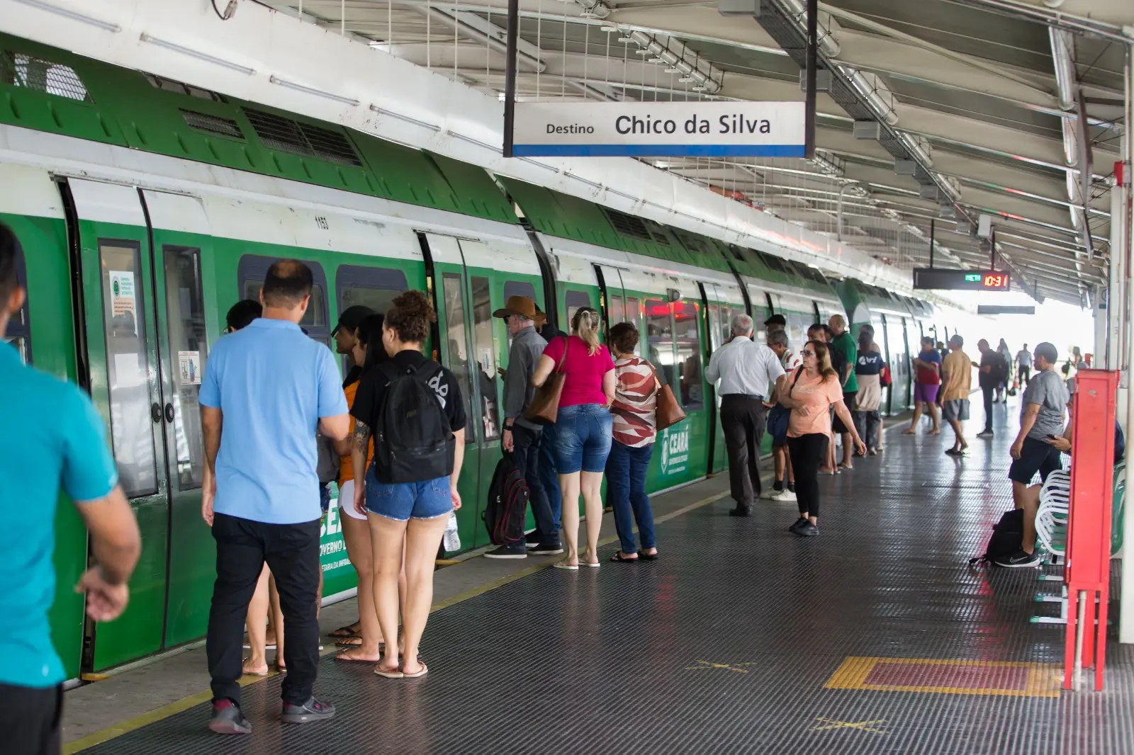 Plataforma de estação do metrô de Fortaleza com passageiros aguardando embarque ao lado de um trem verde parado. Uma placa suspensa indica o destino “Chico da Silva”, enquanto pessoas caminham, esperam em fila e algumas estão sentadas ao fundo da estação coberta.
