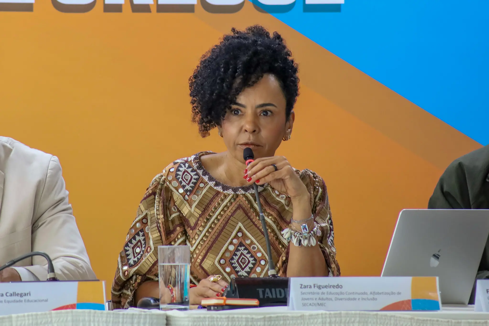 Close em Zara Figueiredo, uma mulher negra de cabelos cacheados e vestindo blusa com motivos africanos, sentada conversando com jornalistas e segurando um microfone de mesa.