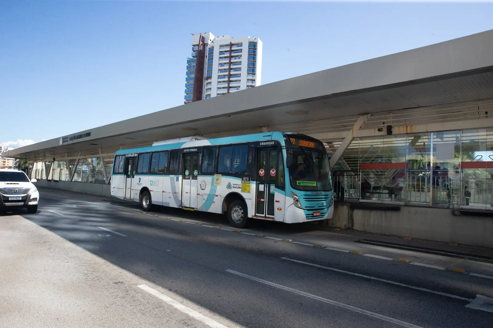 Fotografia em ângulo aberto mostra ônibus urbano de Fortaleza na linha Siqueira parado em estação de vidro e aço do BRT Aguanambi. A estrutura da estação cobre o veículo. Em segundo plano, destaca-se um edifício moderno sob o céu claro.