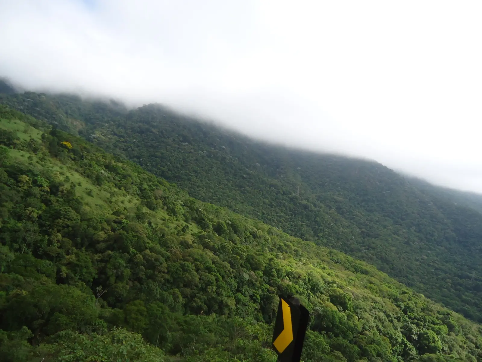 Montanhas cobertas de vegetação verde e densa névoa ao fundo, com uma placa amarela de estrada em primeiro plano, vista da lateral da via em região montanhosa.
