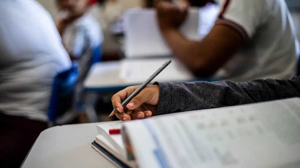 Em primeiro plano, detalhe da mão de um estudante segurando um lápis sobre um caderno aberto em uma sala de aula. Ao fundo, outros alunos aparecem desfocados.
