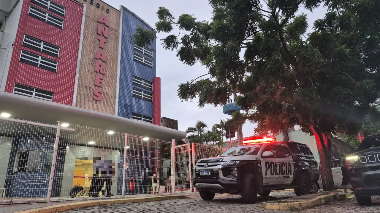 Viatura da Polícia Militar estacionada em frente ao Colégio Antares, no bairro Papicu, em Fortaleza, ao anoitecer. A fachada colorida da escola aparece ao fundo, enquanto agentes e algumas pessoas estão na entrada do prédio.
