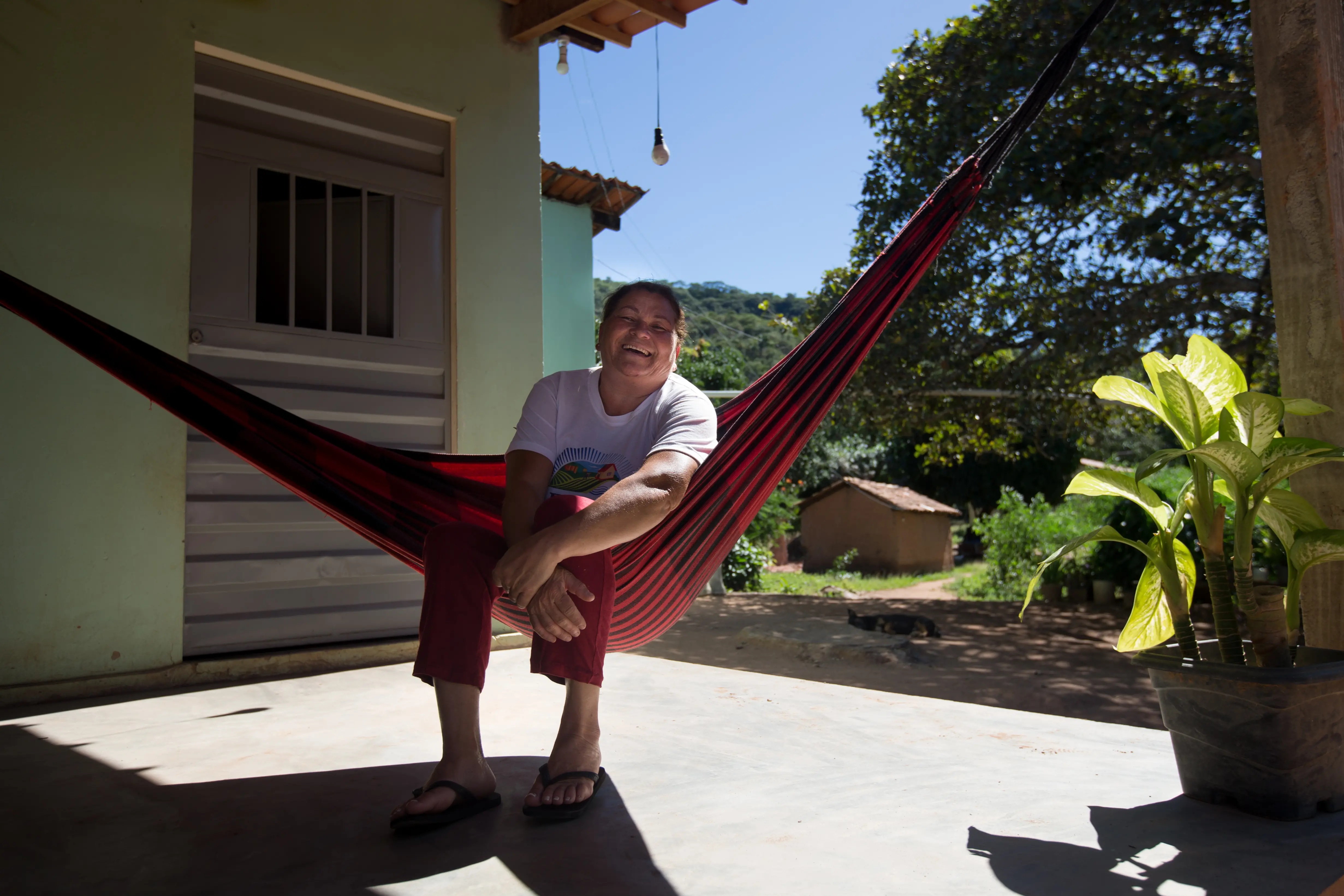 Na imagem, Uma mulher de meia-idade (Dona Celi) sorri alegremente sentada de lado em uma rede de descanso listrada em tons de vermelho e preto. Ela está na varanda de uma casa de campo, com piso de cimento, vestindo uma camiseta branca, calça comprida avermelhada e chinelos. Ao fundo, vê-se um quintal ensolarado com árvores, uma pequena construção de barro (casa de taipa) e um céu azul. À direita, em primeiro plano na varanda, há um vaso de plantas com grandes folhas verdes e amarelas. A iluminação natural cria sombras marcadas no chão da varanda.