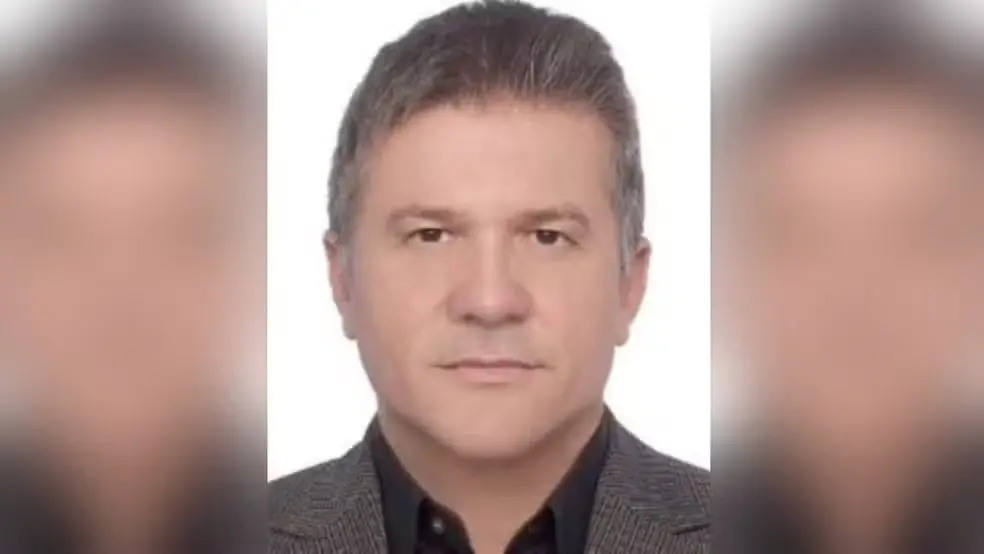 Foto de retrato de um homem, com cabelo grisalho e olhar sério, em fundo branco, usando camisa escura e blazer cinza. Imagem tipo documento.