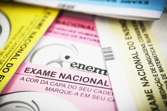Cadernos de prova do Exame Nacional do Ensino Médio (ENEM) aparecem sobrepostos em um plano detalhe, com destaque para a capa de cor rosa que traz o logotipo do exame em preto no centro. As bordas da imagem revelam partes de outros cadernos nas cores amarelo, azul e cinza-claro, mantendo um foco suave que enfatiza os textos institucionais das avaliações.
