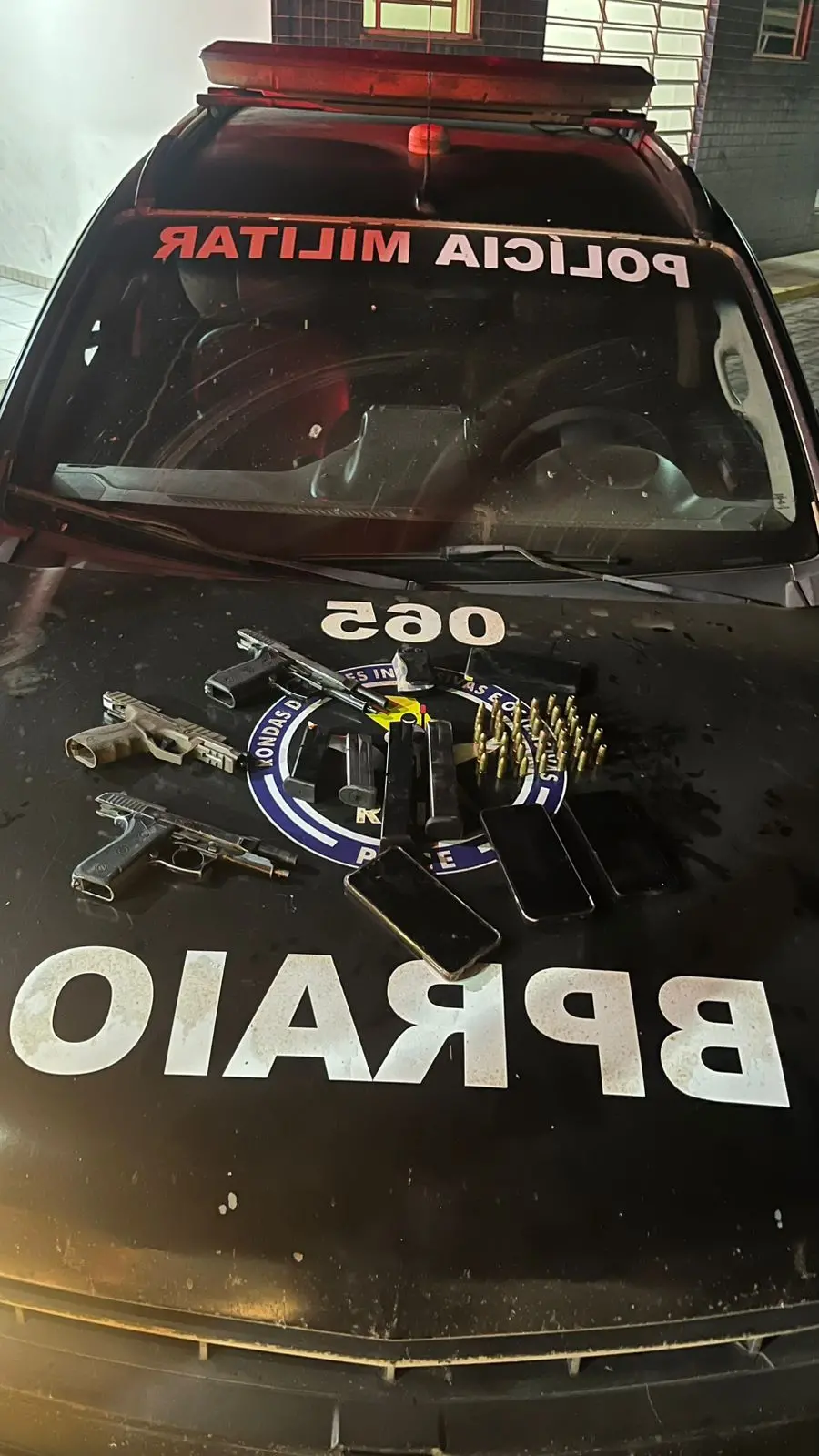 Parte frontal de um carro blindado, com o texto “POLICIA MILITAR” no capô e várias armas e munições posicionadas sobre o painel, em cenário de operação