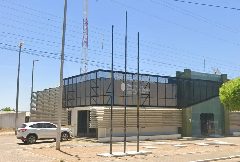A fachada da Delegacia Municipal da Polícia Civil de Aurora, no Ceará, apresenta uma estrutura moderna com painéis escuros na parte superior e brises horizontais brancos nas janelas. Diante do prédio térreo, destacam-se três mastros de bandeira vazios fixados no chão de terra, enquanto um carro SUV branco aparece estacionado à esquerda sob o céu azul e ensolarado.