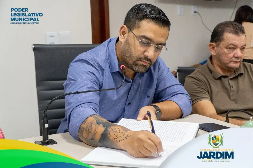 Homem de barba e cabelo curto, usando camisa clara, fala ao microfone enquanto lê em um púlpito durante uma sessão oficial. Ao fundo, há bandeiras — incluindo a do Brasil — e um ambiente institucional. No canto inferior esquerdo, aparece o texto “Sessão Extraordinária – Transição de Cargo”, e no canto direito, o logotipo da Câmara do Município de Jardim com um horário exibido no topo da imagem.
