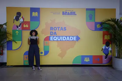 Mulher posa em frente a um painel amarelo com a frase “Rotas da Equidade”, em evento do Ministério da Educação e do Governo Federal. O cenário tem ilustrações coloridas com símbolos de diversidade, educação e inclusão social.