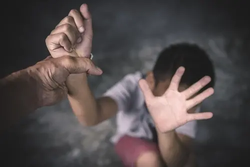 Fotografia em plano detalhado com foco seletivo que retrata uma situação de violência ou abuso. No canto inferior esquerdo, em primeiro plano e com nitidez, a mão forte de um adulto segura firmemente pelo pulso o braço esquerdo de uma criança. Ao fundo, ligeiramente desfoada, a criança — que aparenta ser um menino de cabelos escuros e veste camiseta branca e bermuda rosa — está encolhida no chão. Ela estende a mão direita aberta em direção à câmera, em um gesto defensivo de 