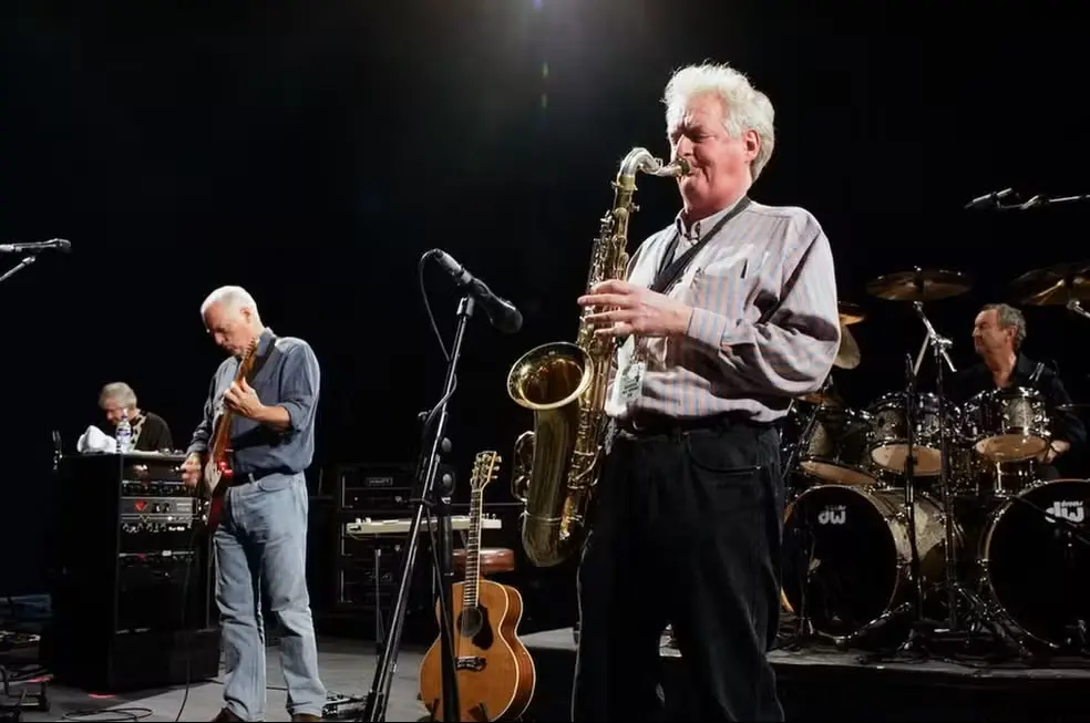 Dick Parry em primeiro plano, em cima do palco, com uma blusa cinza e calça preta, tocando o saxofone. Em segundo plano, um guitarrista tocando guitarra e um homem tocando bateria.