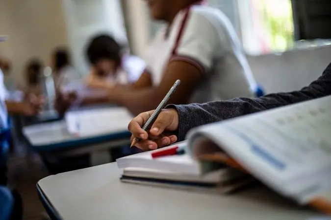 Mão de criança com lápis, escrevendo em livro, em sala de aula. Ao fundo, silhuetas de outros alunos.