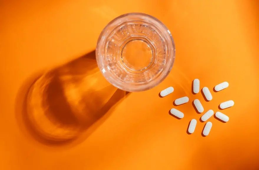 Copo com líquido transparente em um fundo laranja. Ao lado do copo, algumas pílulas de remédio espalhadas