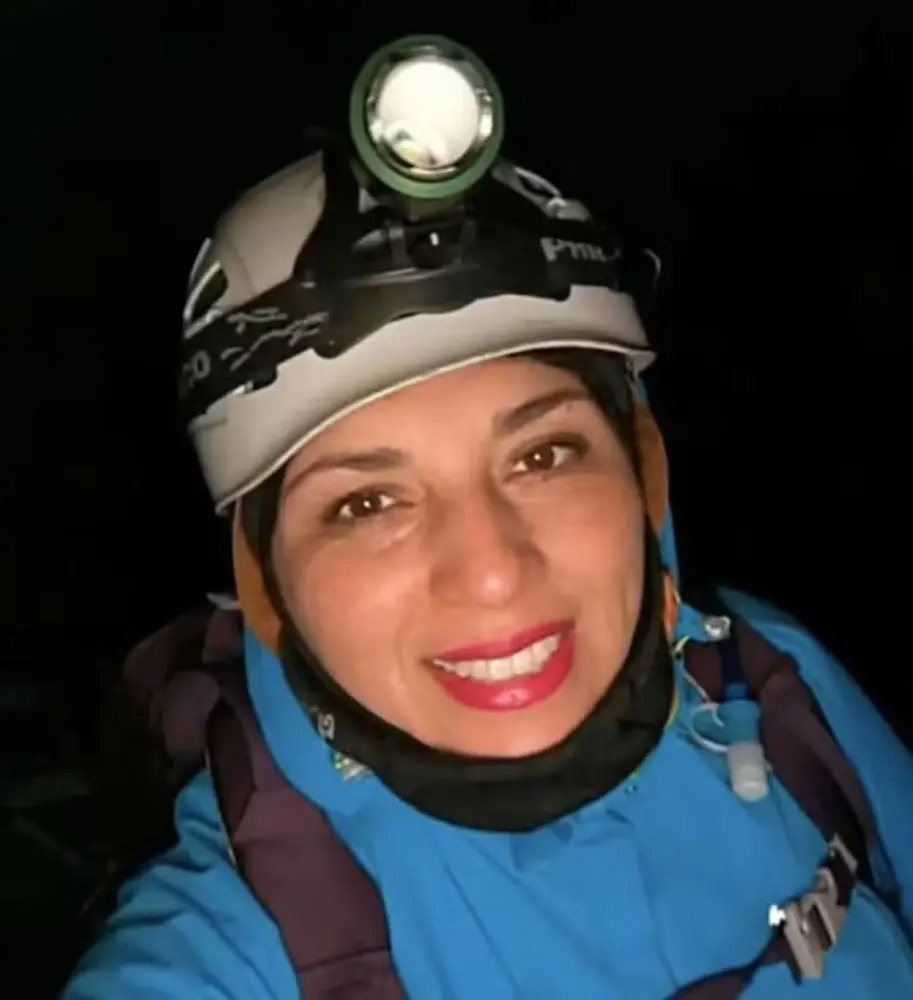 Na imagem, selfie de uma mulher sorrindo, identificada como a alpinista Ingrid, contra um fundo totalmente escuro. Ela tem pele clara, olhos castanhos e usa batom vermelho destacado. Está equipada para o montanhismo, vestindo uma jaqueta azul de frio com capuz sobre a cabeça, uma proteção preta no pescoço e alças de uma mochila nas costas. Na cabeça, ela usa um capacete de proteção cinza claro com uma lanterna de cabeça acesa e direcionada para a frente.