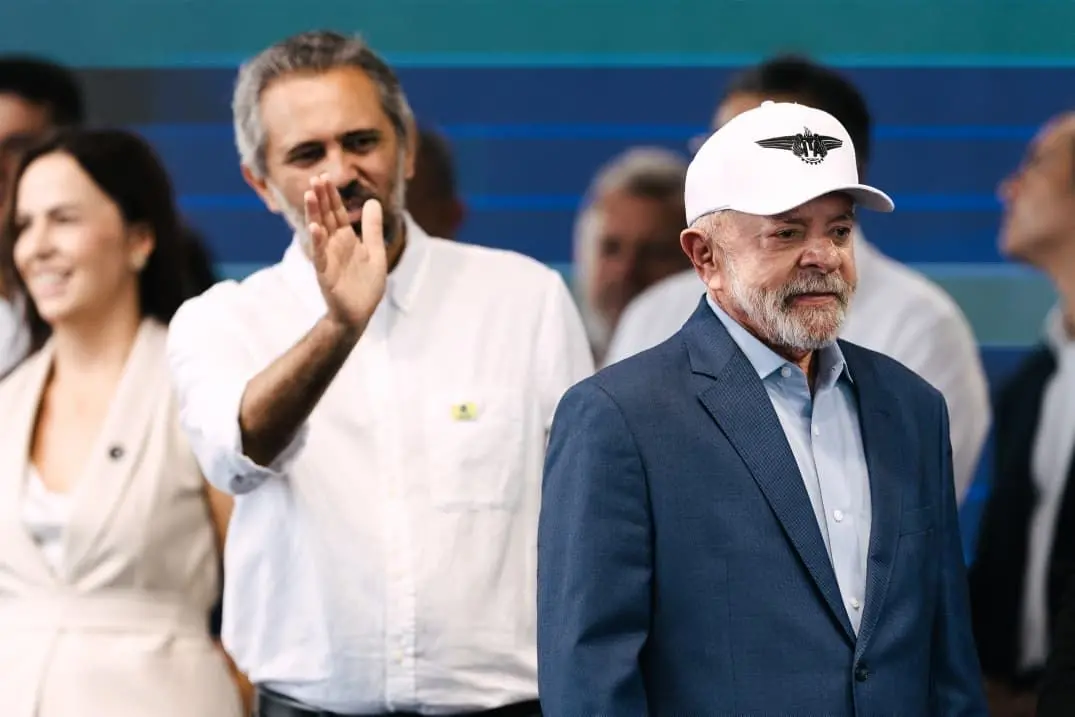 O presidente Lula aparece de terno azul e boné branco durante evento, acompanhado do governador Elmano de Freitas ao fundo, que acena para o público. Outras pessoas participam da agenda ao redor.