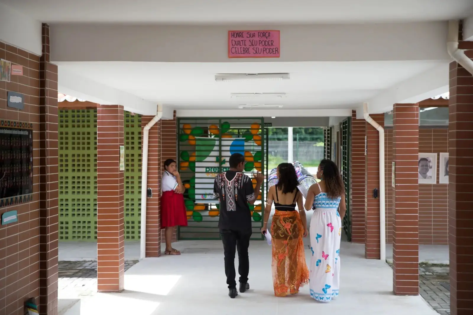 Corredor de escola indígena com estudantes caminhando em direção à entrada principal. Três jovens aparecem ao centro usando roupas com elementos culturais e estampas coloridas. Ao fundo, há um painel verde e amarelo na grade da escola, enquanto uma estudante observa encostada em uma coluna. Na parte superior do corredor, um cartaz traz a frase: “Honre sua força, exalte seu poder, celebre seu poder”.