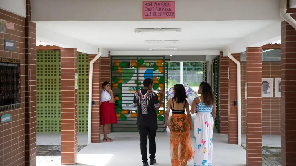 Corredor de escola indígena com estudantes caminhando em direção à entrada principal. Três jovens aparecem ao centro usando roupas com elementos culturais e estampas coloridas. Ao fundo, há um painel verde e amarelo na grade da escola, enquanto uma estudante observa encostada em uma coluna. Na parte superior do corredor, um cartaz traz a frase: “Honre sua força, exalte seu poder, celebre seu poder”.