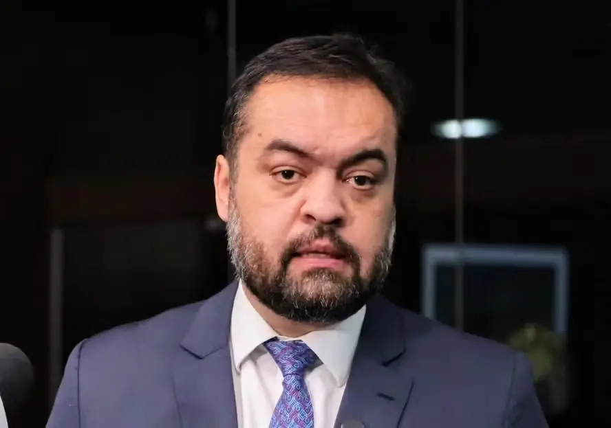 Homem de terno azul e gravata azul com padrão em entrevista, falando em frente a um fundo escuro com microfone à esquerda e iluminação de estúdio.