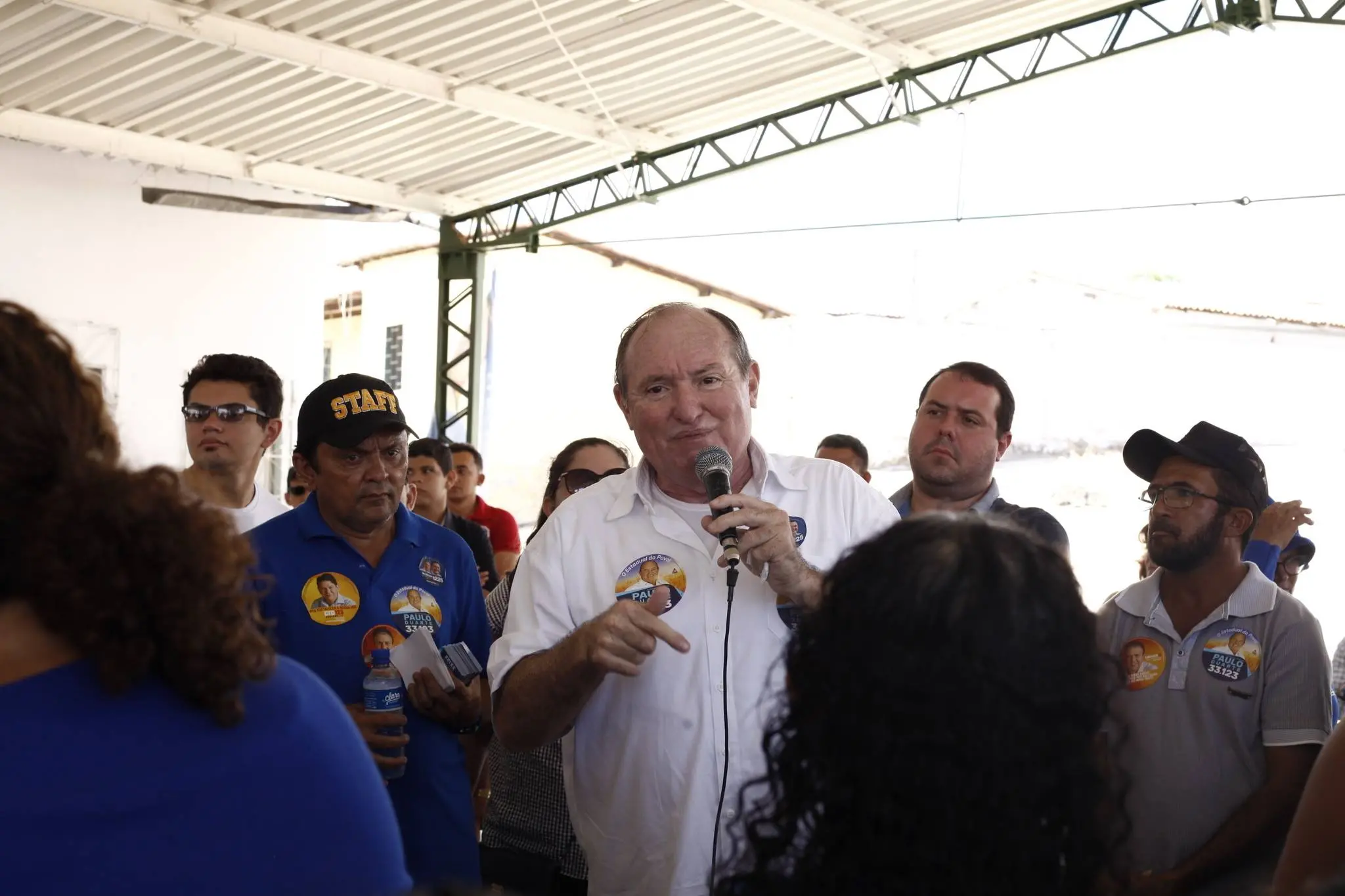 O político Paulo Duarte fala ao microfone durante encontro com apoiadores em espaço coberto. Pessoas ao redor usam adesivos de campanha e acompanham o discurso.