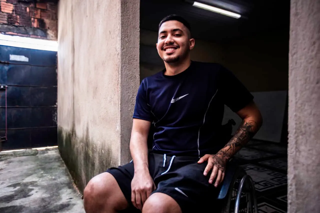Nicolas Henrique, homem de blusa preta e calção preto, sentado em uma cadeira de rodas sorrindo. Ele está na porta de um cômodo da casa, com paredes de cimento e chão de cimento.