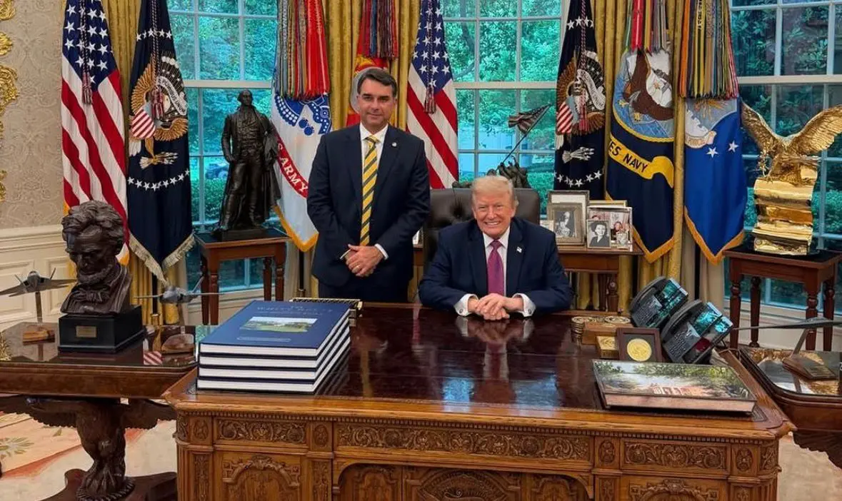 Flávio Bolsonaro posa em pé ao lado de Donald Trump, que está sentado em seu escritório oficial na Casa Branca.
