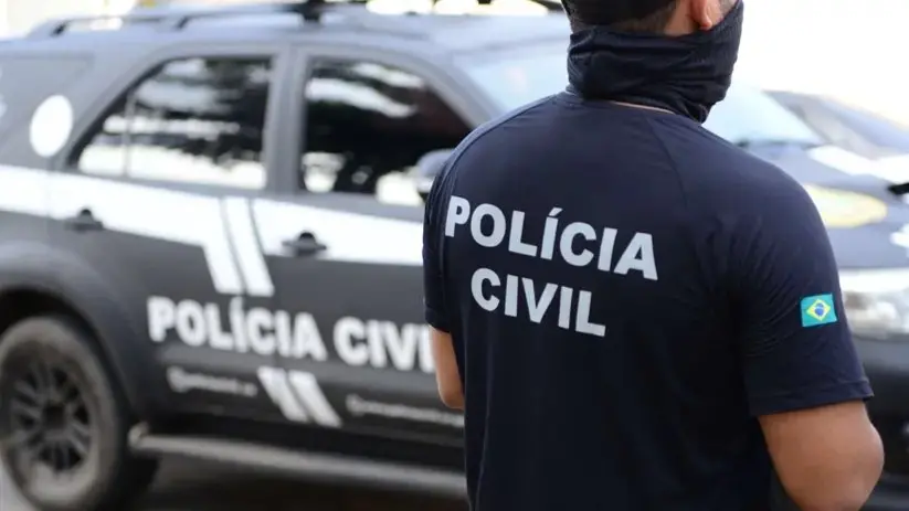 Policial civil de costas, usando uniforme preto com a inscrição “POLÍCIA CIVIL”, ao lado de uma viatura da Polícia Civil em ambiente externo.