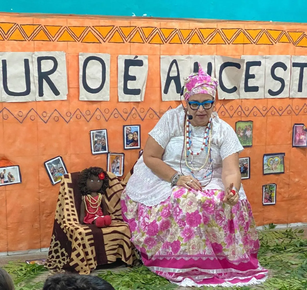 Mulher sentada em frente a um mural laranja com fotos de pessoas negras. Ela está vestida de roupas tradicionais afro-brasileiras. Ao seu lado, uma boneca negra está sentada em uma cadeira menor.