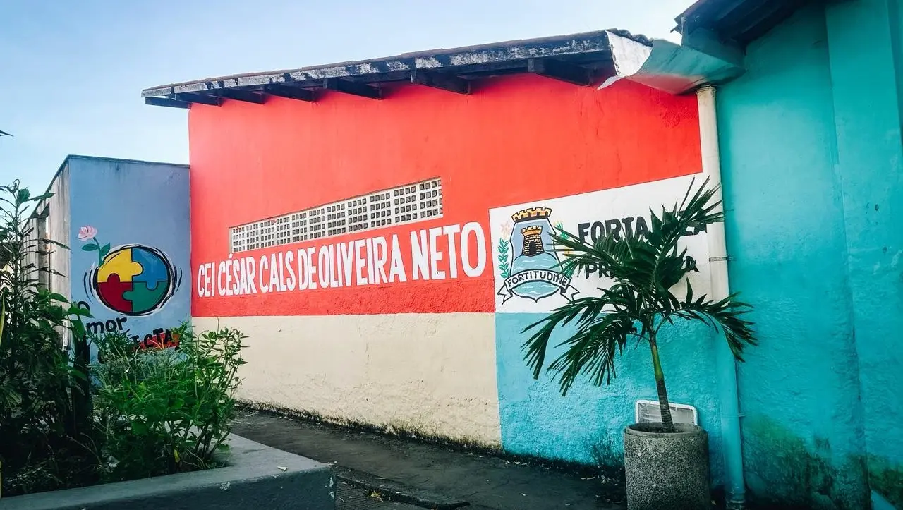 Fachada do Centro de Educação Infantil (CEI) César Cals de Oliveira Neto, em Fortaleza. Muro colorido com o nome da escola e o brasão de armas do município.