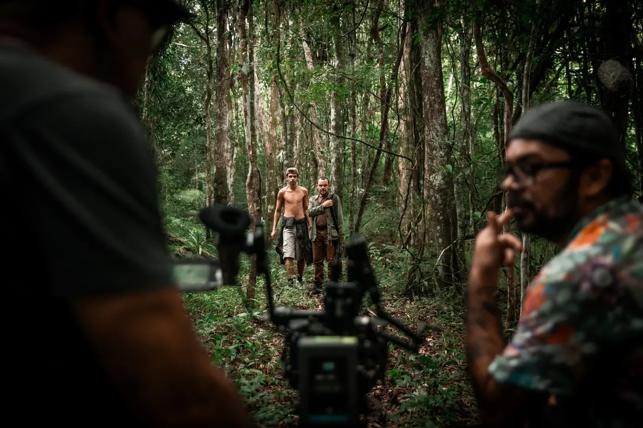 Atores Lucas Galvino e Matheus Nachtergaele caminham entre árvores densas em uma floresta fechada, observados em primeiro plano por uma câmera de cinema e dois membros da equipe técnica de costas e de perfil. A vegetação rústica compõe uma cena de bastidores com tons verdes e terrosos.