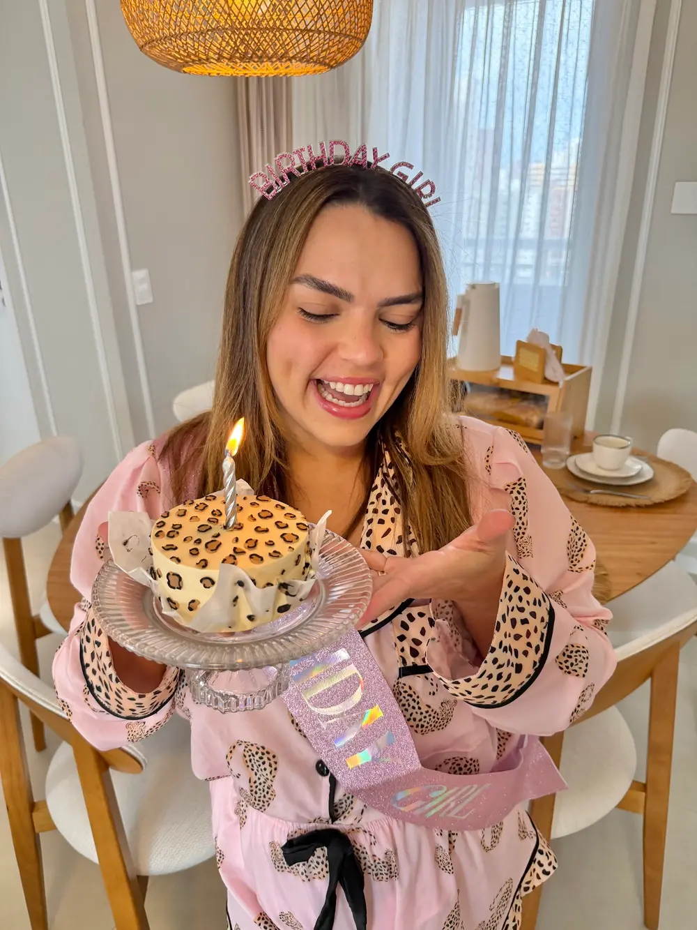 Na imagem, uma mulher jovem, de pele clara e cabelos castanhos claros, sorri alegremente de olhos semi-cerrados enquanto segura um pequeno bolo de aniversário. Ela usa uma tiara rosa brilhante que diz 