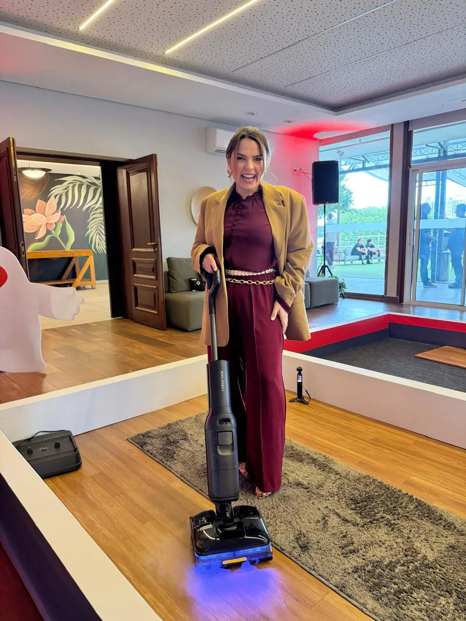 Na imagem, uma mulher jovem, de pele clara e sorrindo alegremente, está de pé no centro de um espaço de demonstração de produtos. Ela veste um macacão longo na cor vinho (burgundy) com gola alta franzida, um cinto de corrente dourada fina e um blazer oversized bege ou caramelo por cima. Ela segura pela alça um aspirador de pó vertical e lavador de piso moderno, na cor preta, da marca Roborock, que projeta uma luz azul em direção ao chão. O aparelho está posicionado sobre uma faixa de tapete cinza felpudo e um piso de madeira clara. A área é delimitada por uma mureta branca baixa. Ao fundo, o ambiente amplo mostra portas de madeira abertas revelando um hall com parede decorada de flores, sofás cinzas e uma grande parede de vidro que dá para uma área externa iluminada.