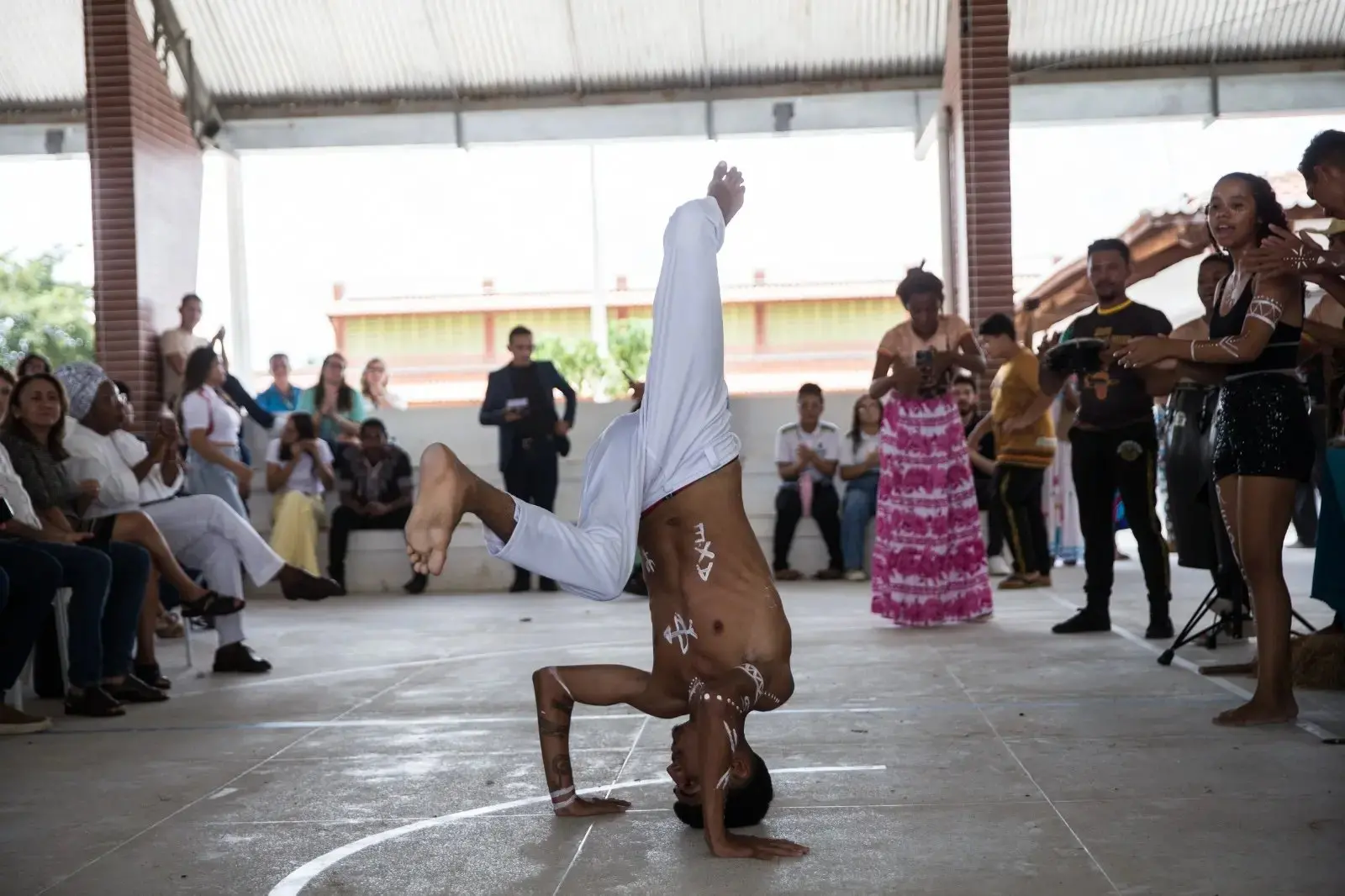 Adolescente negro vestido com calça branca e sem camisa, na quadra de uma escola, fazendo uma apresentação de capoeira.
