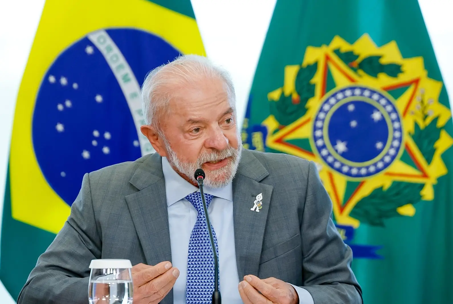 Lula é um homem idoso, branco, de cabelo e barba também brancos. Na foto, ele está gesticulando enquanto discursa, com a bandeira do Brasil atrás. Usa terno cinza, camisa social da mesma cor e gravata azul.
