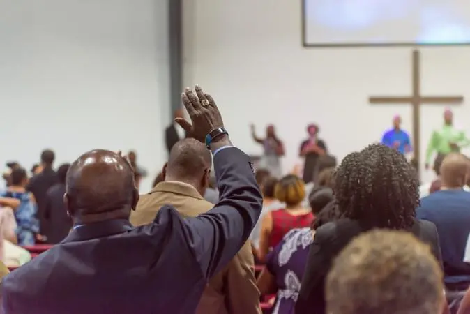 Fiéis participam de culto em igreja, com um homem em primeiro plano levantando a mão durante momento de adoração diante do altar e de uma cruz ao fundo.