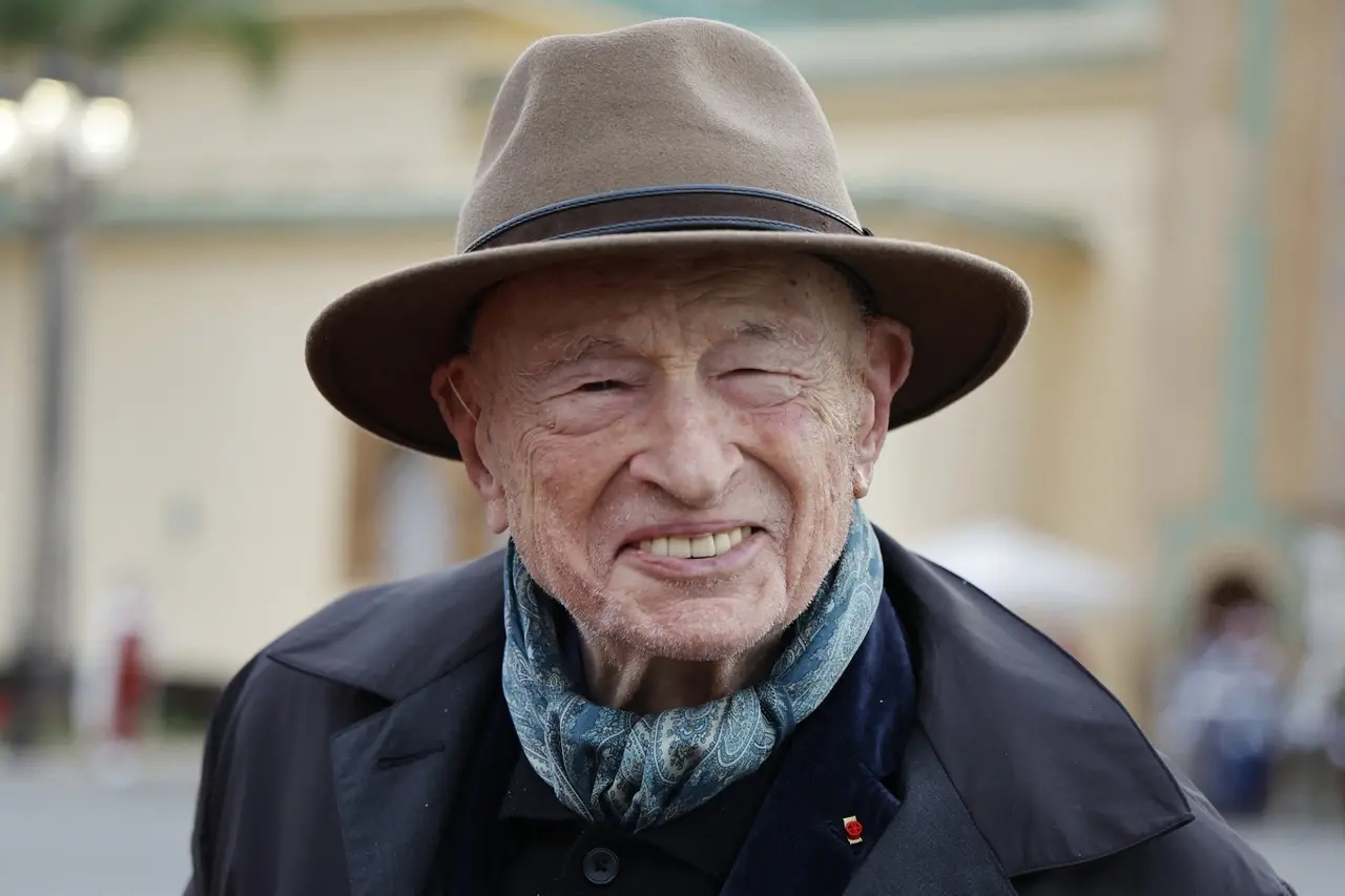 Homem branco idoso, com chapéu bege e sobretudo azul, sorrindo para a câmera.