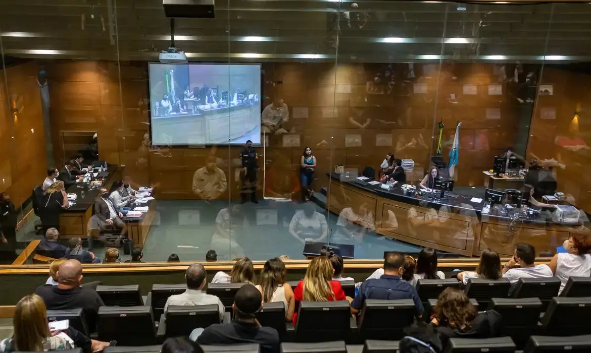 Foto de uma sala de visualização de julgamentos no 2º Tribunal do Júri, no centro do Rio de Janeiro. Sala tem parede de vidro que dá para assistir a sessão. Contém cadeiras com pessoas sentadas.