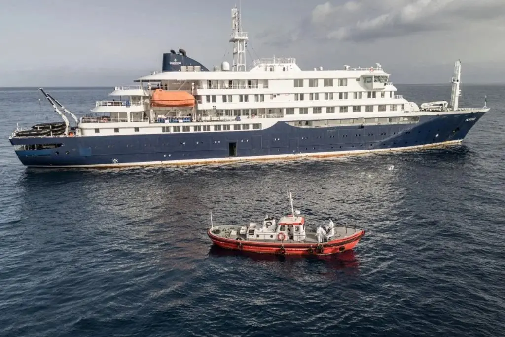 Na imagem, vista aérea de um grande navio de cruzeiro e expedição, o MV Hondius, navegando em um mar de águas escuras sob um céu parcialmente nublado. O navio possui o casco azul-escuro na parte inferior e vários andares com cabines brancas na parte superior, com botes salva-vidas laranjas acoplados à lateral. Em primeiro plano, uma embarcação menor de apoio, com casco vermelho e cabine branca, navega paralelamente ao navio de grande porte. É possível ver pessoas a bordo do barco menor vestindo trajes de proteção brancos.