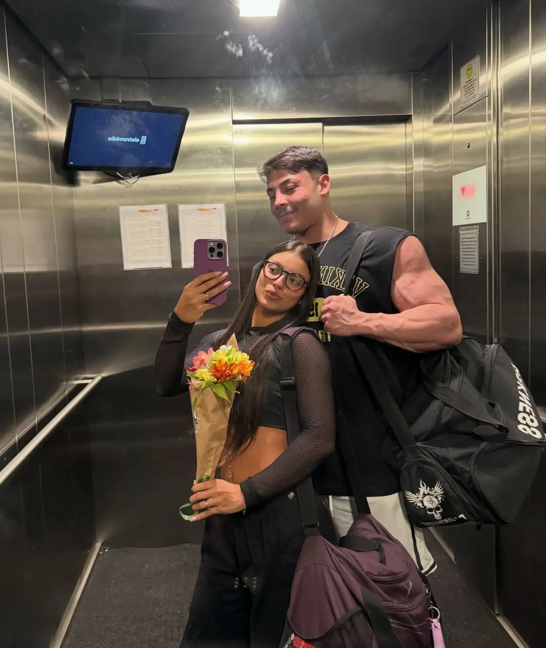 Um jovem casal posa para uma selfie no espelho dentro de um elevador de metal, destacando um homem muito musculoso que flexiona o bíceps enquanto a mulher ao seu lado segura um buquê de flores e registra o momento com o celular.