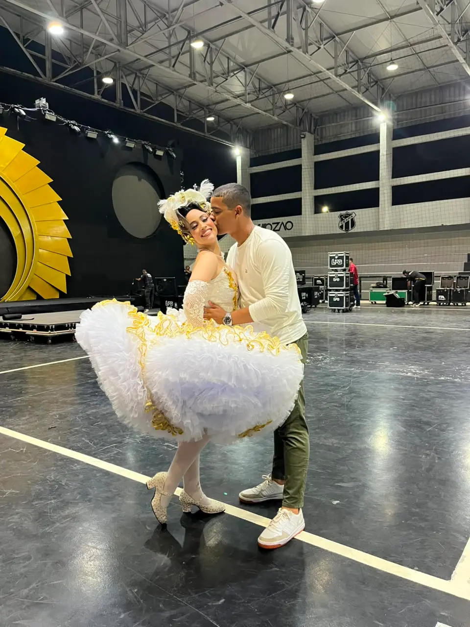 Na imagem, foto de corpo inteiro em plano médio de um casal em um grande ginásio coberto. No centro, uma mulher sorridente está inclinada para trás, usando um traje típico de noiva de quadrilha junina: um vestido branco muito volumoso, cheio de camadas de babados rodados e bordas com fitas douradas, luvas de renda branca e sapatos de salto baixo brancos com texturas. Ela usa um arranjo de cabeça branco com plumas. Um homem jovem, de cabelo curto, veste uma camiseta de manga longa branca, calça verde-oliva e tênis claros. Ele está em pé, abraçando-a pela cintura e dando um beijo carinhoso em sua bochecha. O chão é uma quadra preta com linhas brancas. Ao fundo, o cenário do ginásio mostra uma grande estrutura amarela em formato de sol ou engrenagem à esquerda, equipamentos de som e luz (cases pretos da LFG) empilhados e, na parede dos fundos, o escudo do Ceará Sporting Club com a palavra 