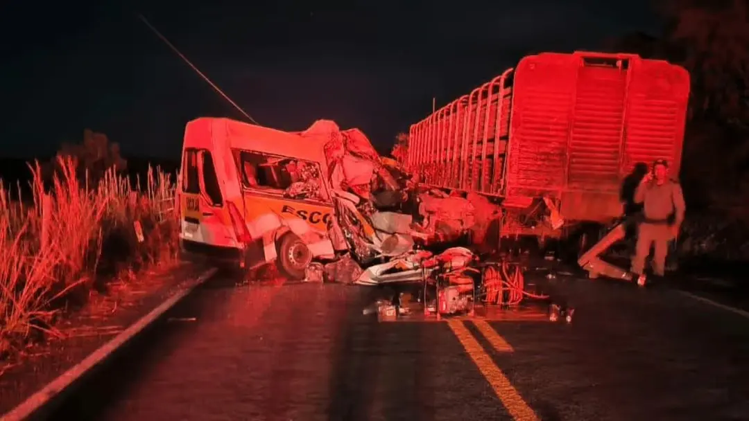 Caminhão e carreta envolvidos em acidente na rodovia, com veículo danificado, detritos e equipes de resgate em meio a fumaça e iluminação vermelha que destaca a cena.